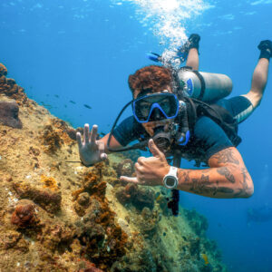 Open Water diver exploring coral reef underwater on scuba, making a hand signal with vibrant marine life in the background.