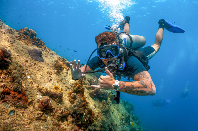 Scuba diver exploring coral reef underwater, making a hand signal with vibrant marine life in the background.