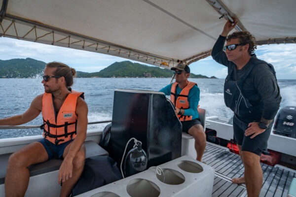 Men on a boat wearing life vests navigate the ocean with scenic island views in the background.