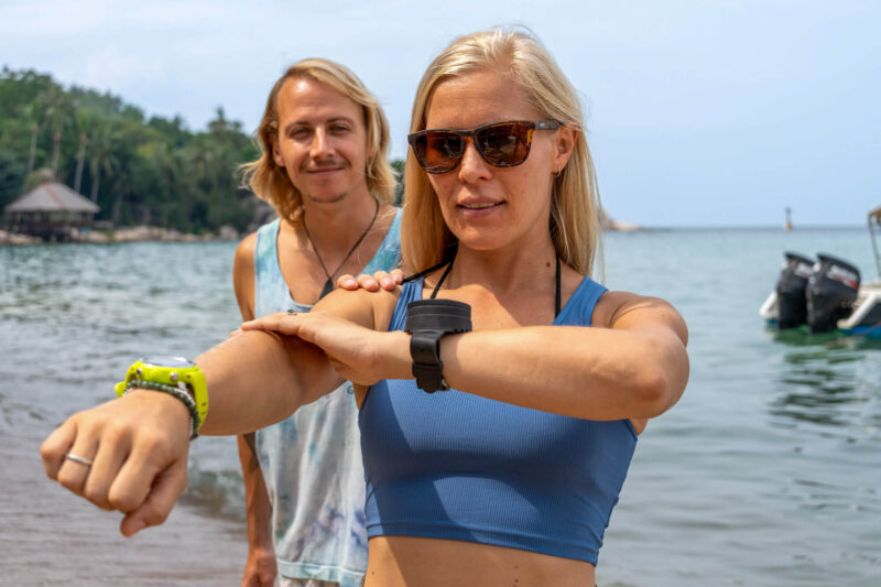Two people preparing for watersports on a tropical beach with boats and palm trees in the background.