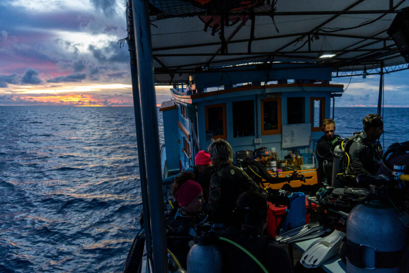 Divers preparing on a boat at sunset, ready for a scuba dive adventure, with the ocean and vibrant skies in the background.