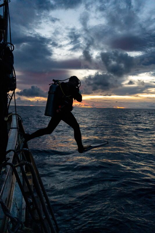 Scuba diver jumping into the ocean at sunset with dramatic clouds in the background.