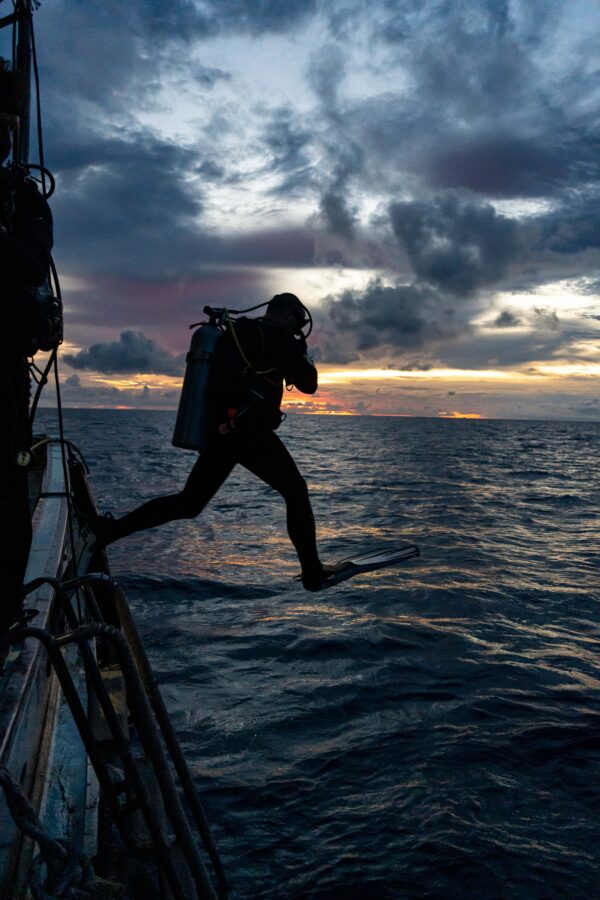 Scuba diver jumping into the ocean at sunset with dramatic clouds in the background.
