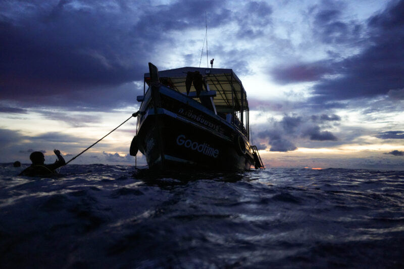 Boat at sea during sunset, silhouetted against a dramatic sky; swimmer in foreground reaching towards the vessel.