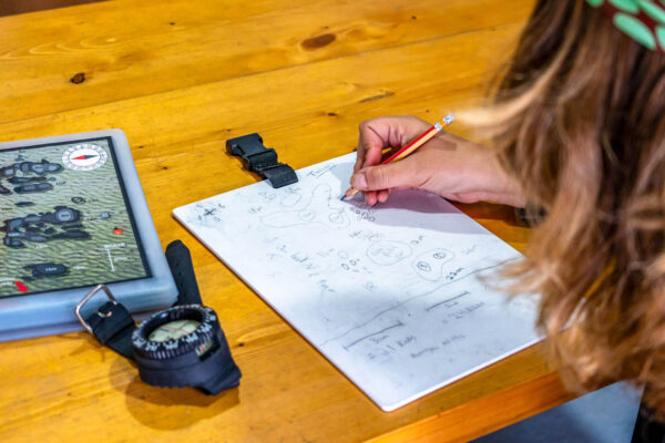 Person sketching a map on paper with a compass and map on a wooden table.