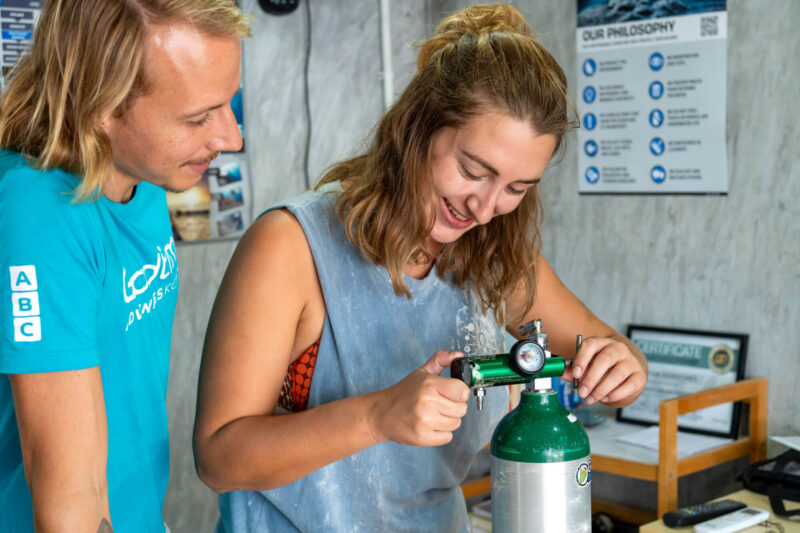Two people learning scuba diving techniques with oxygen tank indoors, smiling and adjusting settings.