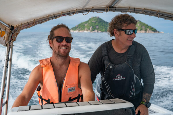 Two men on a boat enjoying a sunny day near a lush island, wearing life jackets and sunglasses.