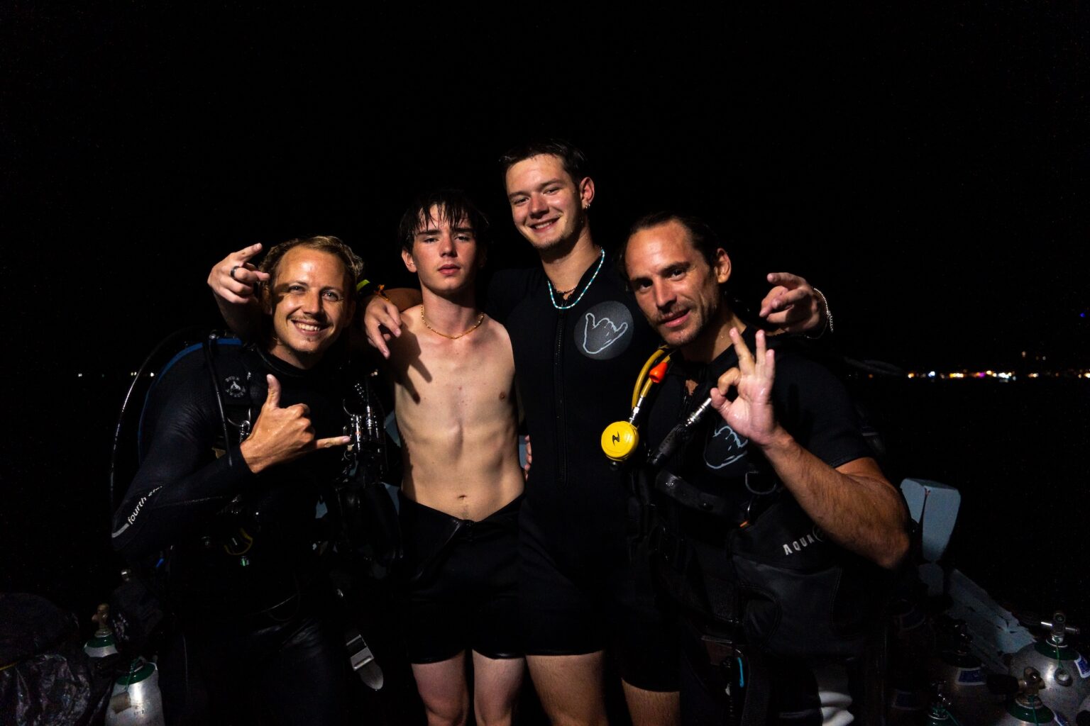 Group of four scuba divers posing cheerfully at night, wearing wetsuits and gear, making hand signs.