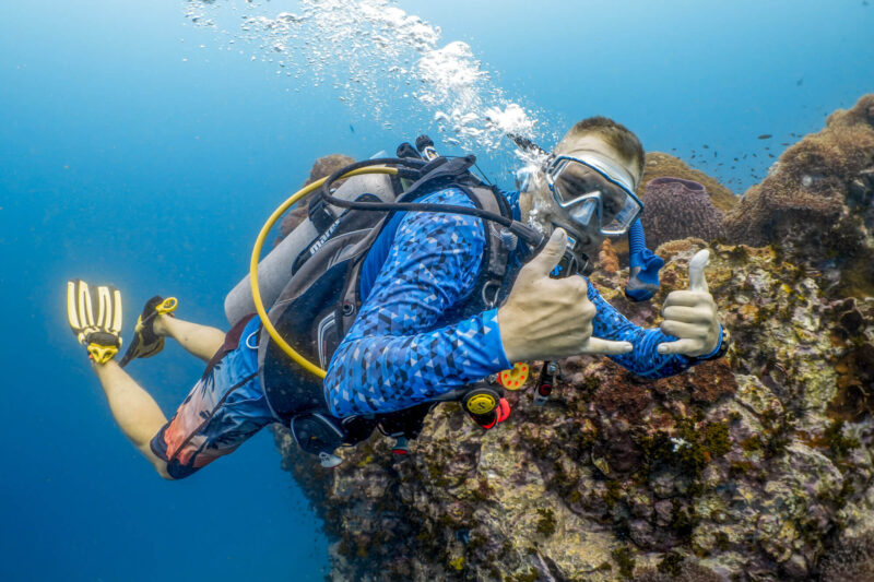Scuba diver exploring vibrant coral reef underwater, showing hand signals with clear blue ocean background.
