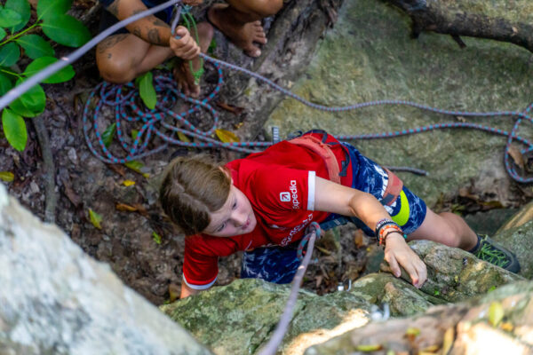 Young climber in red shirt scaling a rocky surface, with ropes and harness, surrounded by a forest landscape.