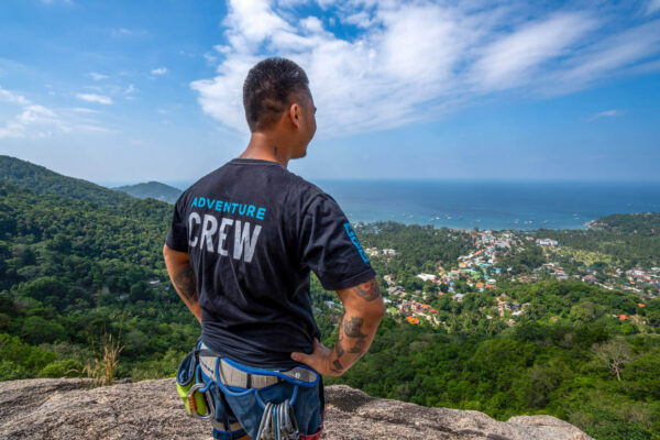 Man in adventure gear overlooks scenic coastal landscape from a cliff, under a bright blue sky.
