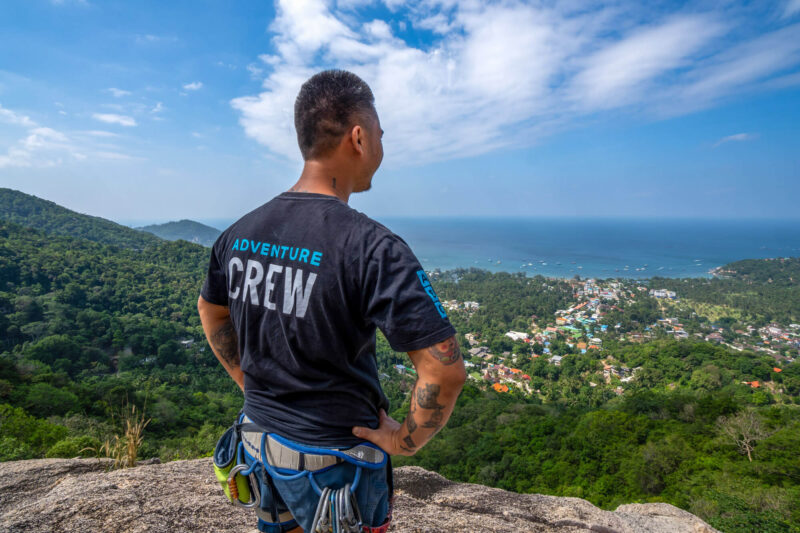 Man in adventure gear overlooks scenic coastal landscape from a cliff, under a bright blue sky.