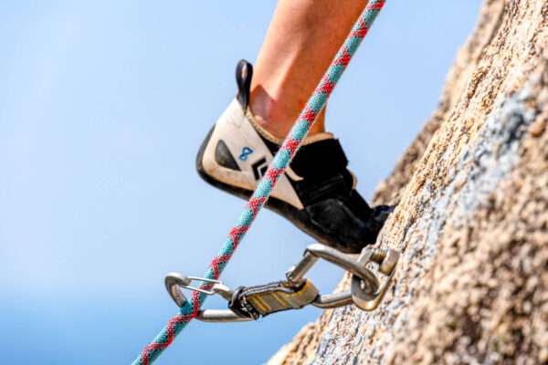 Close-up of climber's foot in shoe on rock, with carabiner and rope ensuring safety during ascent.