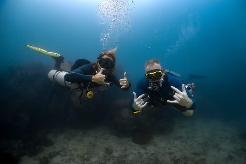 Two scuba divers underwater giving thumbs-up and hand signals, surrounded by coral reef in clear ocean water.