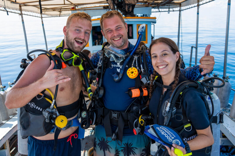 Three divers smiling on a boat, wearing scuba gear and ready for an adventure in clear blue waters.