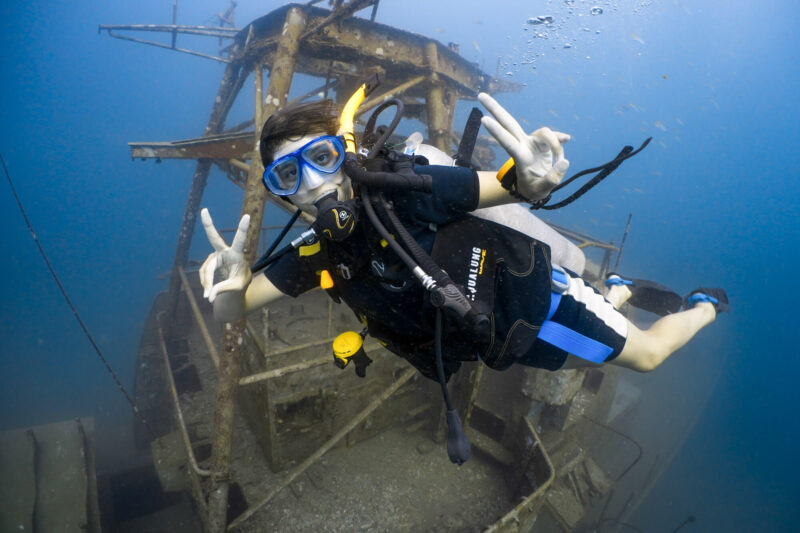 Scuba diver explores shipwreck underwater, posing with peace signs, wearing blue gear.