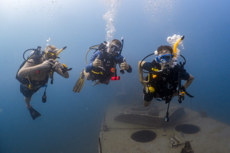 Three scuba divers exploring a shipwreck underwater, displaying hand signals, with bubbles rising above them while scuba diving in Koh Tao Thailand.