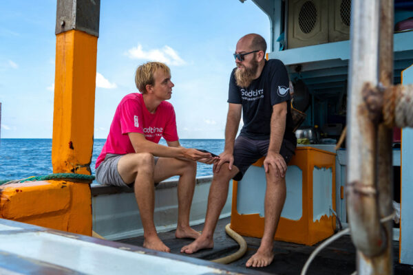 Two men talking aboard a boat on a sunny day, with the ocean in the background.