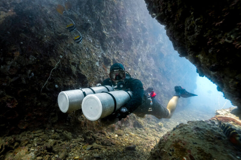 Scuba diver explores underwater cave with dual tanks, surrounded by marine life, in clear ocean water.