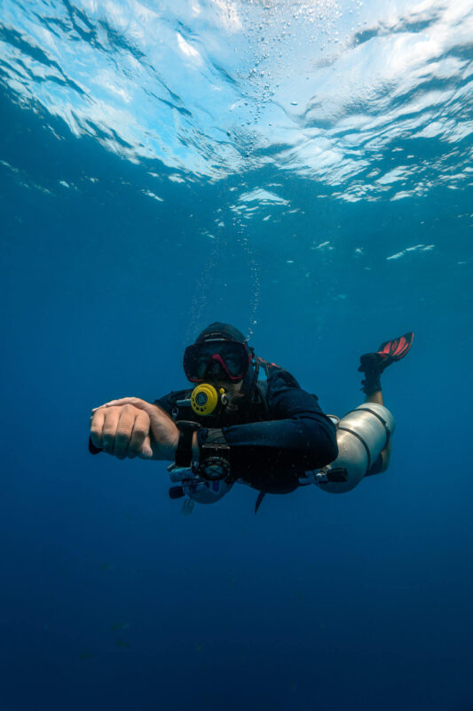 Scuba diver exploring underwater, wearing full gear, with clear blue ocean and sunlight filtering through above.