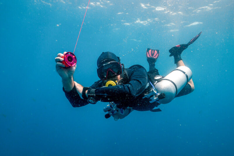 Scuba diver underwater holding a reel, wearing fins and scuba gear, in clear blue ocean depths.
