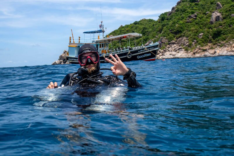 Scuba diver signaling OK in blue ocean with boat and rocky shoreline in the background.