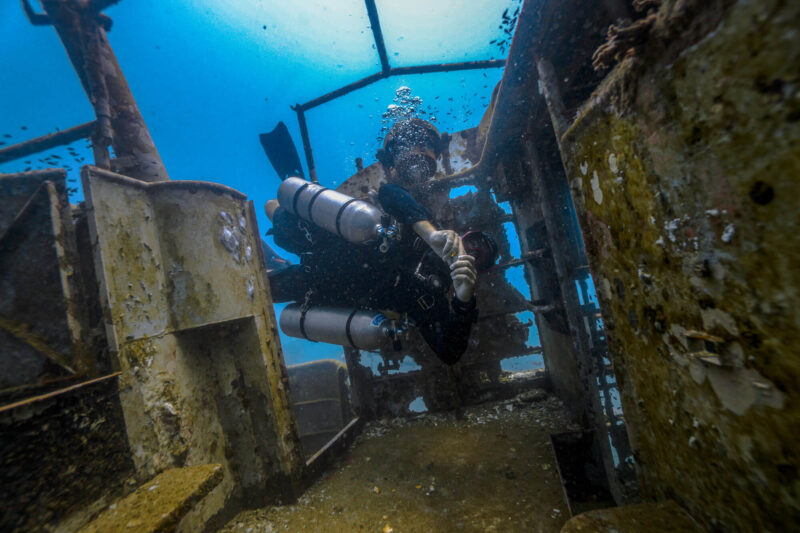 Scuba diver exploring an underwater shipwreck, surrounded by marine life and bubbles.