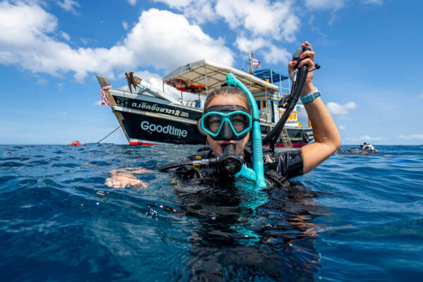 Diver surfaces near boat Goodtime in clear blue ocean, holding snorkel, under a sunny sky.